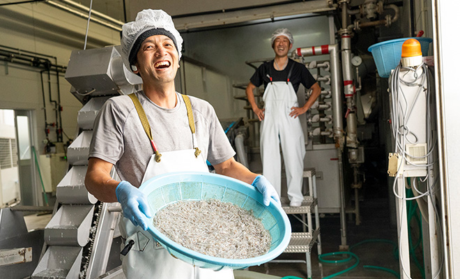 生しらすを手に笑う田中友之さん。後ろでは工場長で社長の息子の森美幸さんがポーズをと