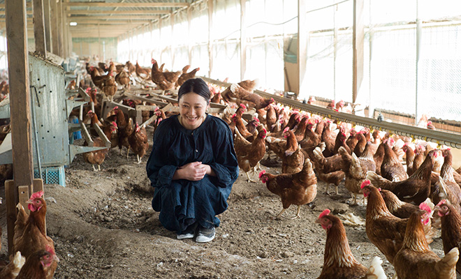 明るい鶏舎で過ごす鶏たちの中にしゃがみ込み微笑む長田佳子さん