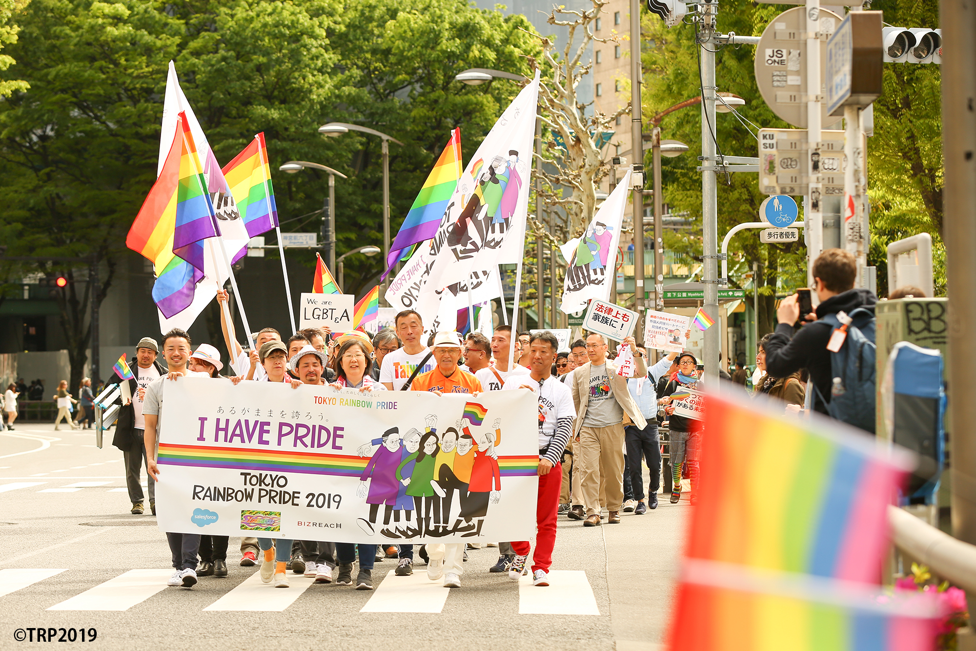7色の虹色の旗や「TOKYO RAINBOW PRIDE」と書かれた横断幕を掲げながら、人々が道路を歩いてパレードする。