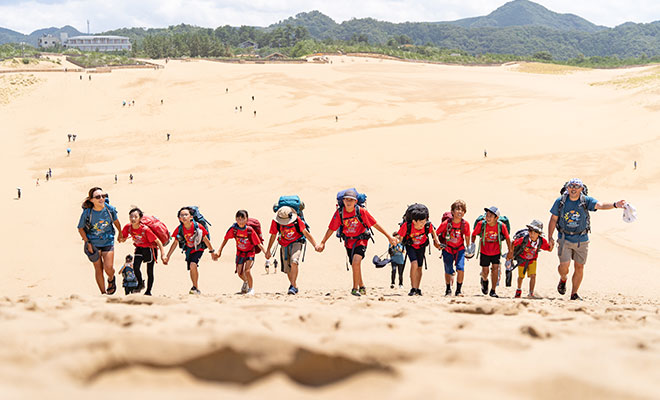 2025年の100マイルアドベンチャーのようす。子どもたりと荻田さんやスタッフが手をつないで砂漠でジャンプしている